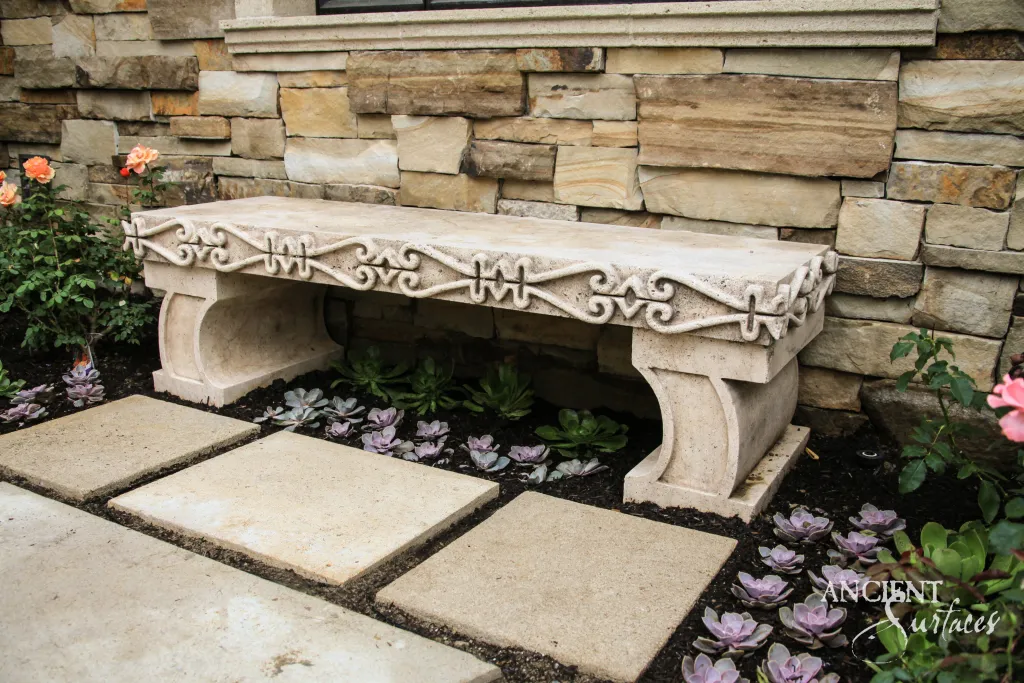 Carved antique limestone garden bench with a scrollwork frieze set against a stacked stone wall