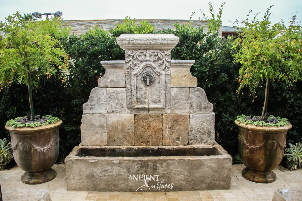 Garden wall fountain assembled from reclaimed limestone blocks, its carved relief panel pouring into a long stone trough