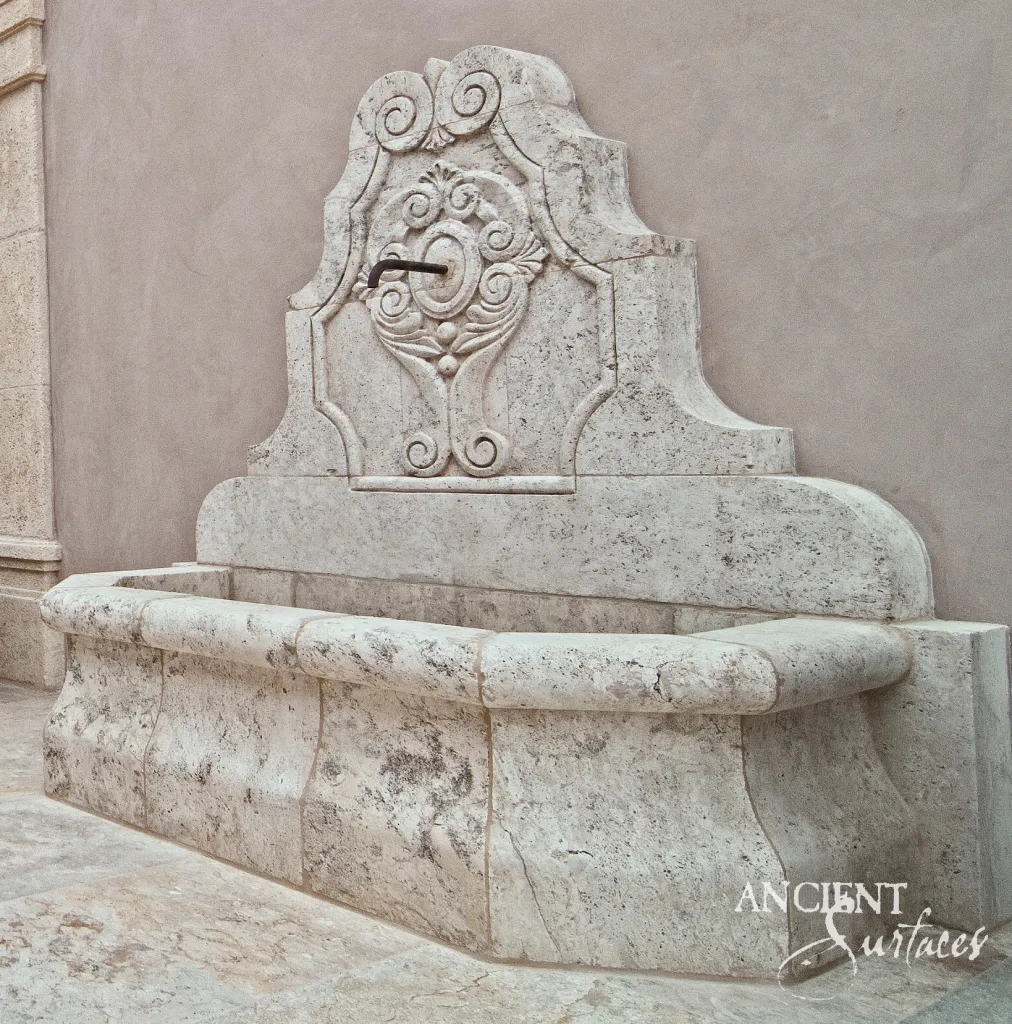 Hand carved limestone wall fountain with a scrolled crest and a curved basin standing on an estate patio